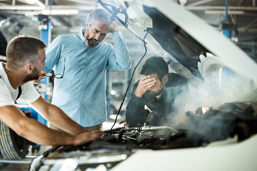Two partners attractive guys, cheerful specialist repairman in blue costume, hat, holds his modern device, businessman in formal classy wear, blurred silver car behind. Vehicle breakdown, check two mechanics explaining to client what is wrong with car